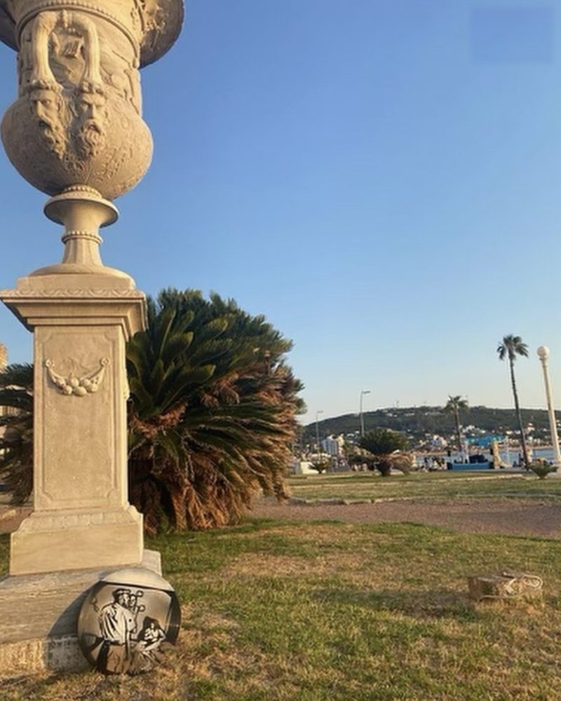 A GEM Movement record piece of Kendrick Lamar at the base of a stone pedestal beside a palm tree on the waterfront, Piriápolis, Uruguay